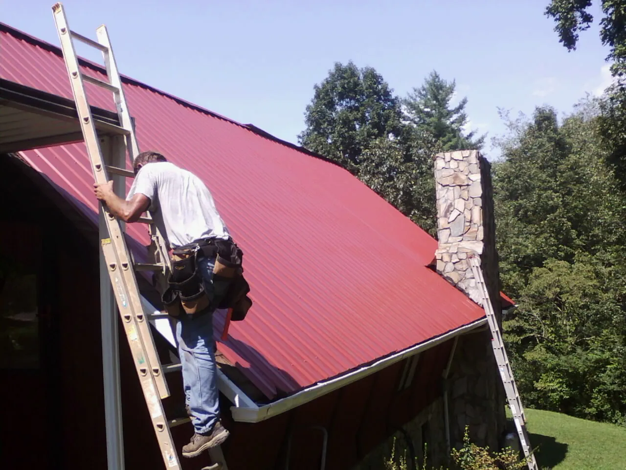 Roofer on a ladder beside a red metal roof and stone chimney in Portage